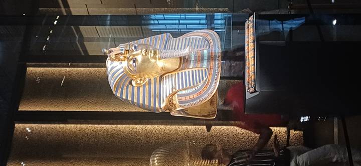 Gold funerary mask of Tutankhamun displayed behind glass inside a dimly lit museum gallery.