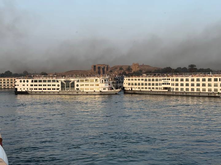 Two large Nile cruise ships docked side-by-side on a hazy morning with Luxor temple ruins in the distance.