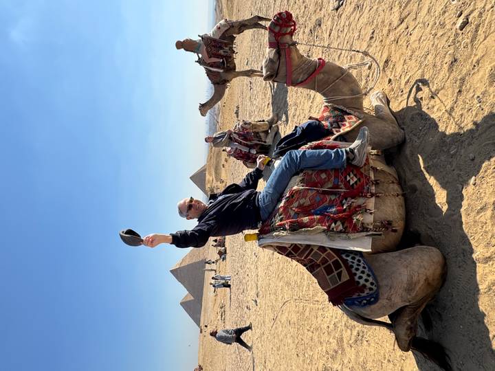 Traveler raising his hat while sitting on a camel with Giza’s pyramids in the sandy background.