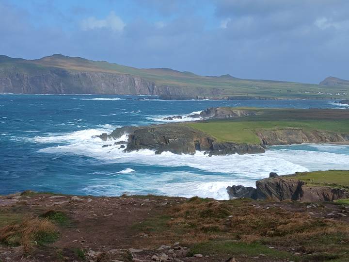 Wild Atlantic waves crash against rugged green headlands beneath a partly sunny sky.