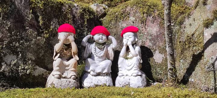 Three small stone monk statues wearing bright red knitted caps enact the ‘see no evil, hear no evil, speak no evil’ poses amid moss-covered rocks.