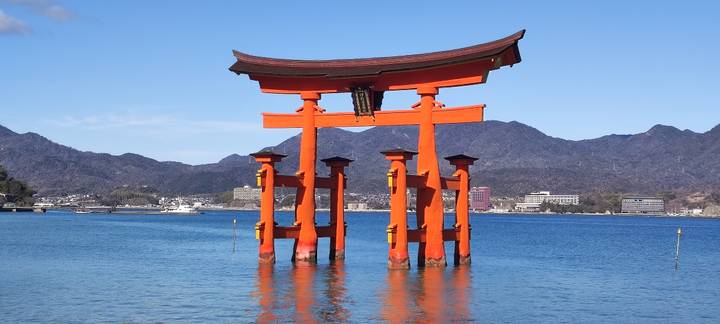 Iconic bright orange torii gate of Itsukushima Shrine stands in calm blue sea backed by rugged mountains and a cloudless sky.