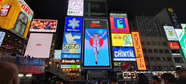 Famous Glico running man sign among many illuminated billboards in busy Dotonbori at night.