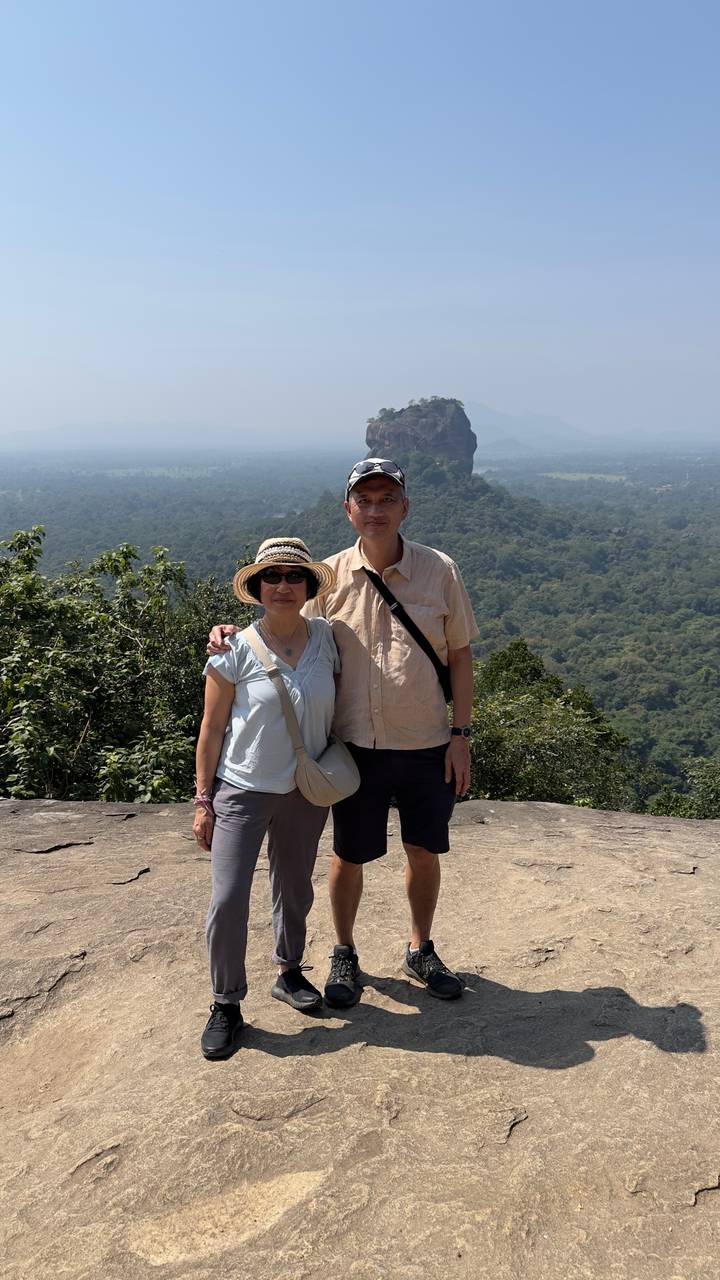 A couple stands arm-in-arm on a rocky outcrop overlooking lush Sri Lankan forest and a distant rock fortress.