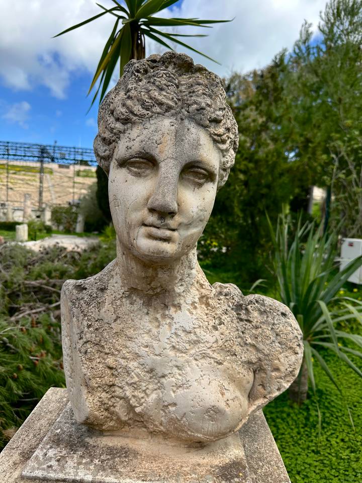 Weathered ancient marble bust statue set in a garden with greenery behind.
