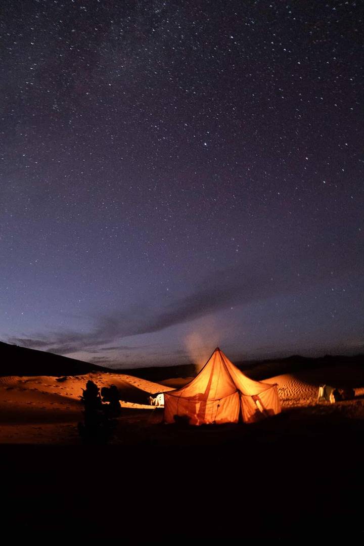 A star-filled desert night sky stretches above a warmly lit triangular tent emitting soft smoke.