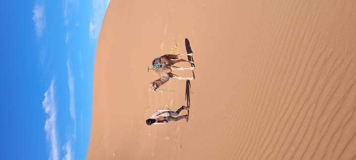 A guide leads a saddled camel across rolling orange dunes under a bright blue sky.