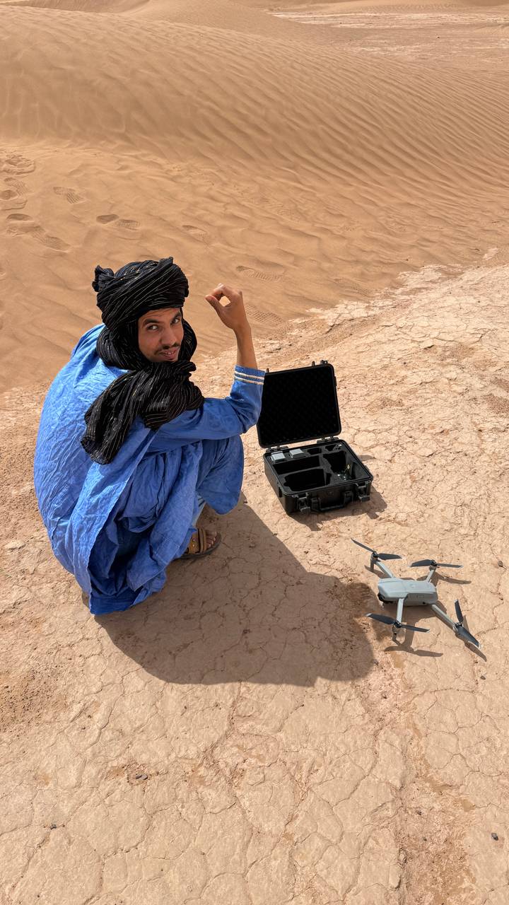 A man in a blue robe and head scarf crouches beside a drone case and small drone on cracked desert ground.
