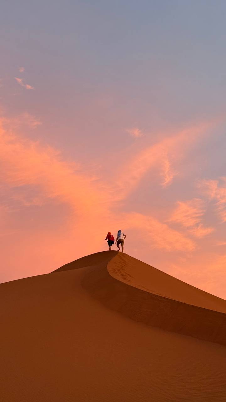 Two small silhouettes climb the crest of a sand dune against a dramatic pink-orange sunset sky.