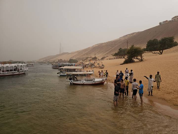 Locals and tourists wading and playing along a sandy Nile riverbank with boats anchored nearby on a hazy afternoon.