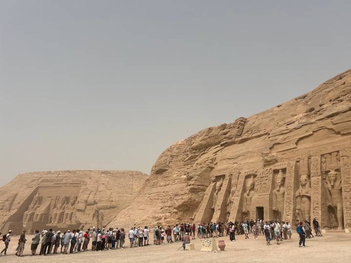 Sweeping view of the colossal rock-cut temples of Abu Simbel with crowds gathered below under a hazy sky.