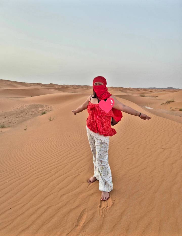 Woman in red scarf spreads her arms joyfully atop endless rolling sand dunes at dusk.