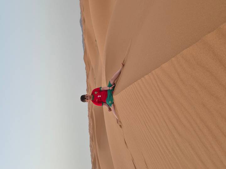 Boy in football jersey sits on the crest of a sand dune at twilight with patterned ridges leading to the horizon.