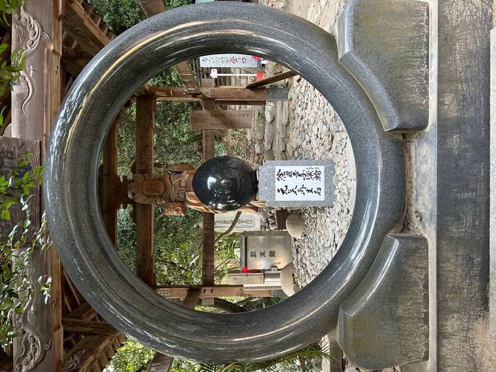 A polished stone ring frames a black sphere and carved wooden statue inside a Shinto shrine courtyard.