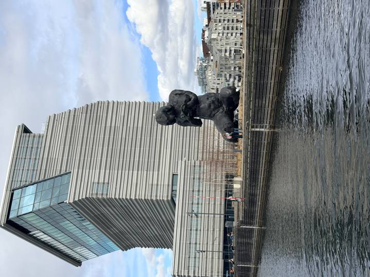 Massive black kneeling sculpture beside modern angular museum building along a waterfront promenade.