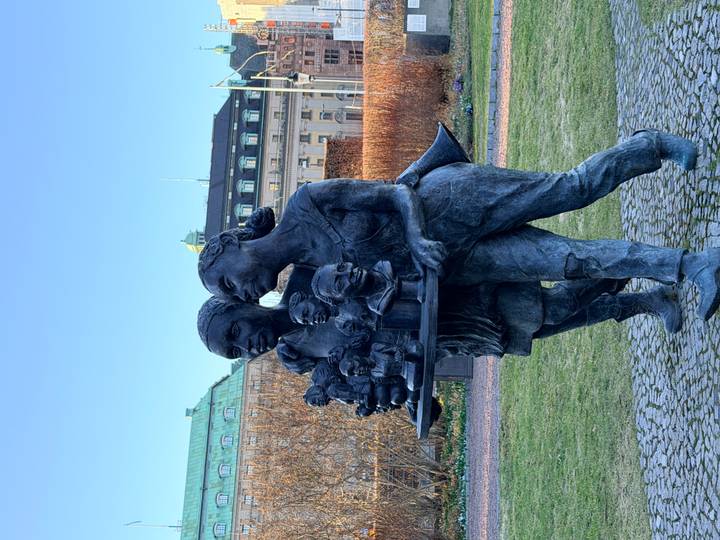 Bronze sculpture of two women carrying small busts through a city park with elegant buildings in the background.