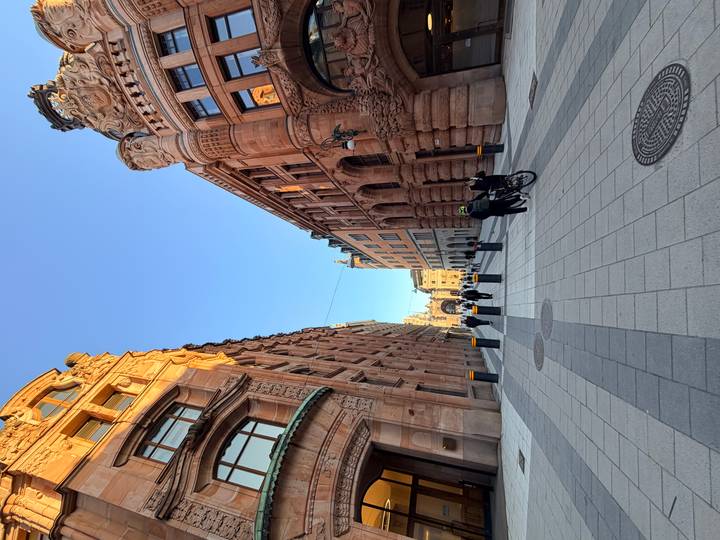 Wide pedestrian street lined with ornate stone buildings and people walking under a clear blue sky