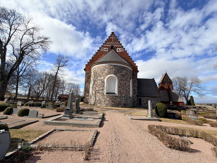 Medieval stone church with patterned brick roof surrounded by old gravestones under patchy clouds