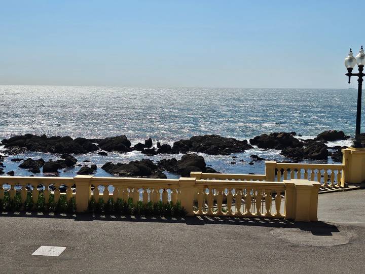 Sunny seaside view with crashing waves and rocky shoreline seen beyond a yellow balustrade promenade.