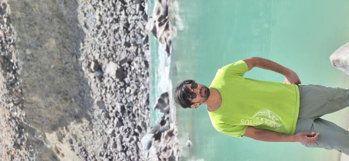 Man in sunglasses stands beside a turquoise mountain river with rocky banks and steep slopes behind.