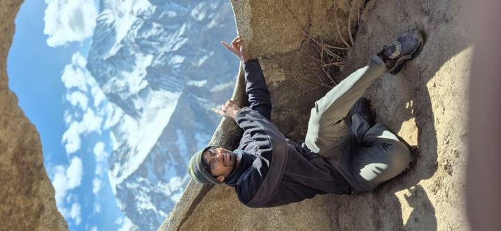 Kneeling hiker points toward towering snow-clad peaks framed by rugged rocks and blue sky.
