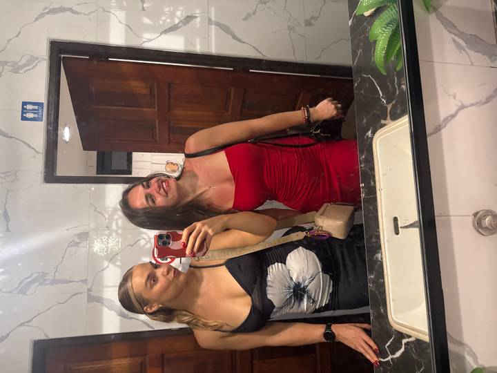 Two young women taking a mirror selfie in a tiled restroom before a night out.
