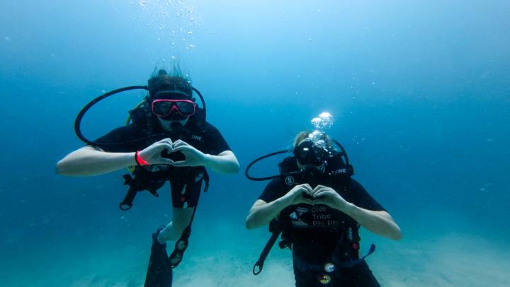 Two scuba divers making heart symbols with their hands in clear blue water.
