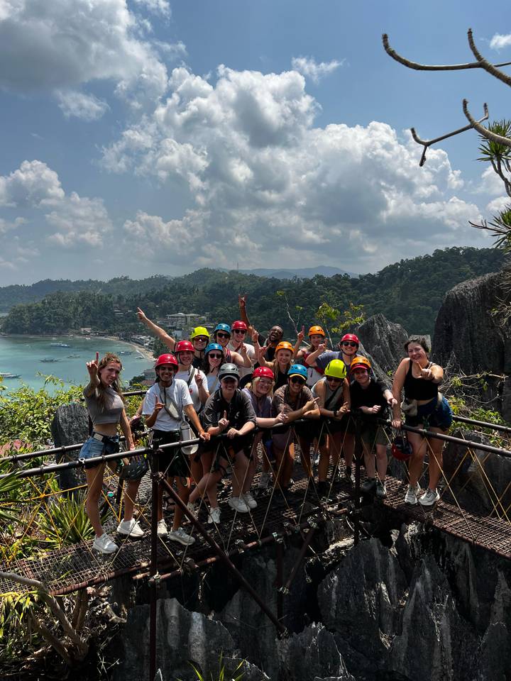 Helmeted adventurers celebrate on a high bamboo bridge with emerald bay and jungle behind.