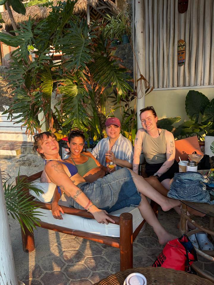 Four women lounge on beachside cushions with drinks during golden late-afternoon light.