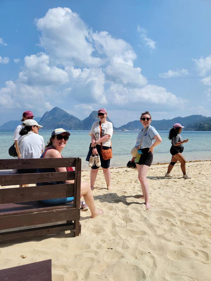 Travellers stand barefoot on sandy island beach with forested karst peaks rising behind them.