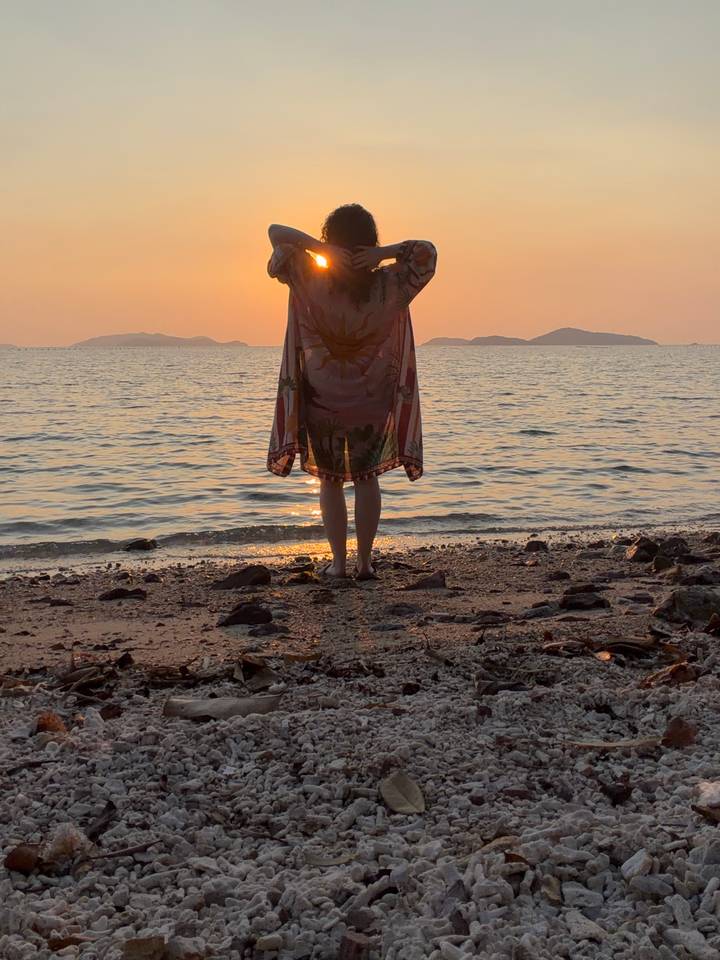 Silhouette of a person wrapped in a shawl standing on a rocky shore during a vivid orange sunset.