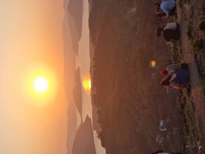 Travellers sit on a ridge watching the sun set over a winding bay and forested hills.