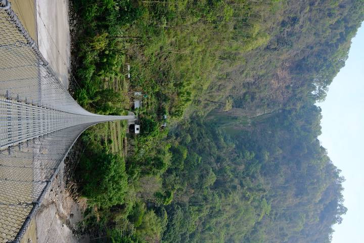 Long suspension bridge stretches over a rocky river toward small houses set against steep green mountains.