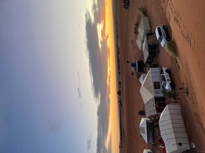 Canvas desert camp with 4x4 vehicles sits beneath a vivid sunset sky over rolling sands.