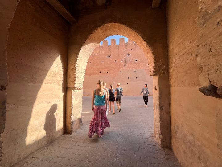 Visitors walk through a sunlit stone arch into a walled courtyard.