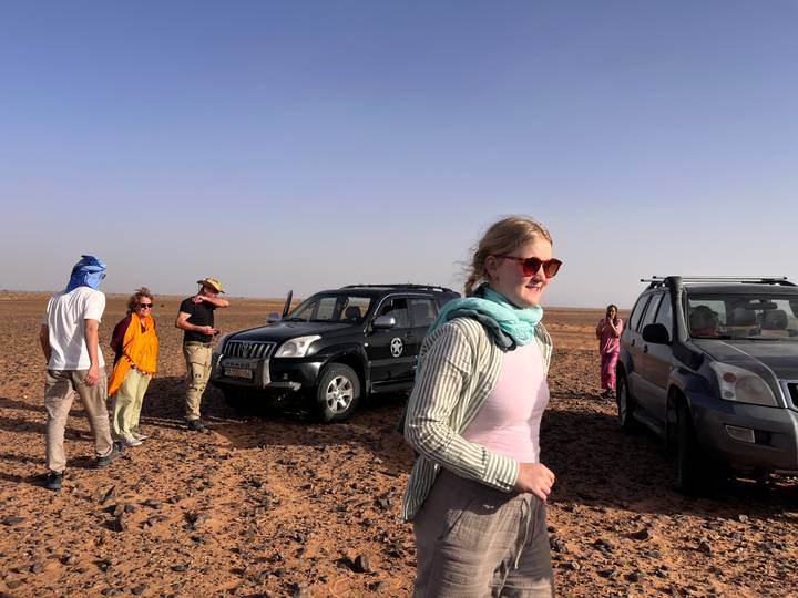 Travellers walk across a dry desert plain near parked SUVs under a clear sky
