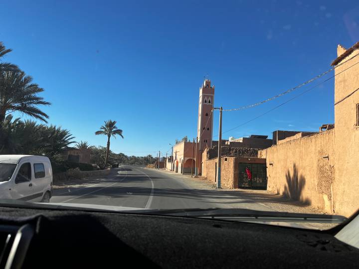 View from vehicle driving through a sunlit desert town with minaret and palm trees