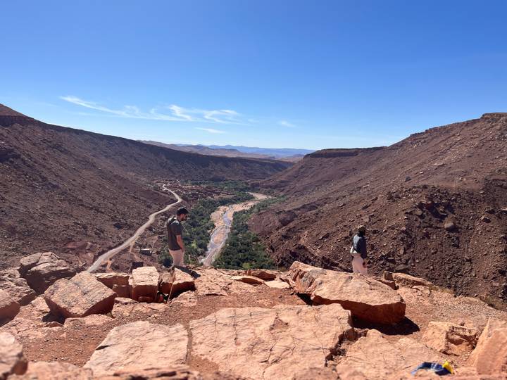 Wide shot of two travellers exploring edge of vast reddish gorge with river below