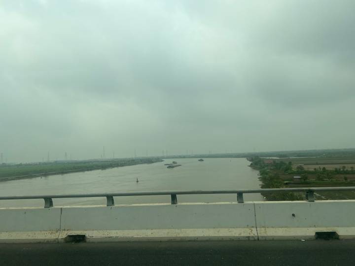 Distant barges navigating a broad brown river on a grey cloudy day.