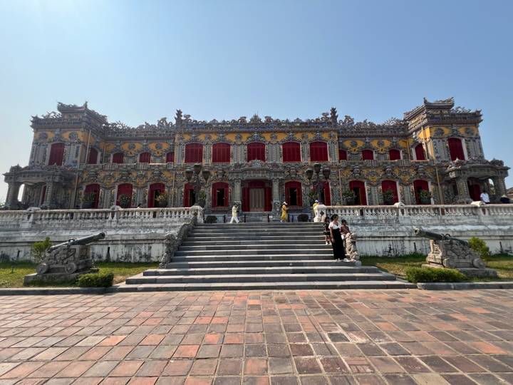 Ornate yellow building with red shutters and cannons, visitors on the grand staircase.