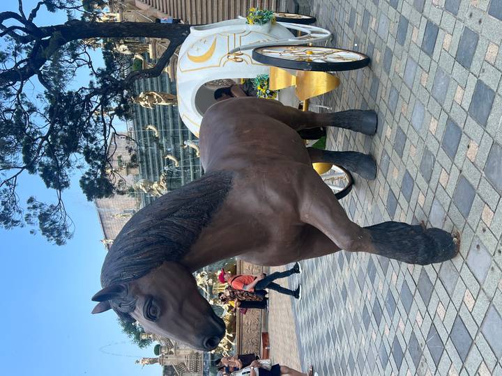 Life-size brown horse statue hitched to a decorative carriage in a public square.