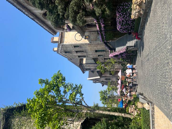 Charming cobblestone street lined with flowering walls and cafés packed with tourists under a clear sky.