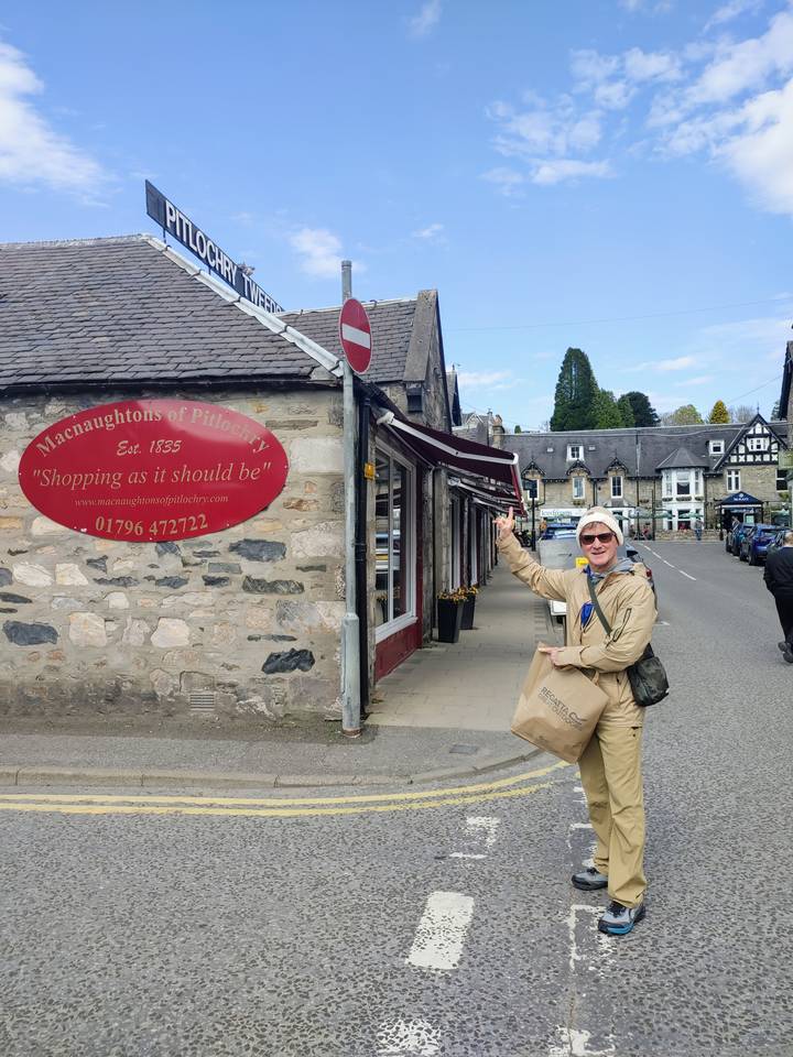 Smiling traveler pointing at a red oval shop sign on a quaint stone street in Pitlochry.
