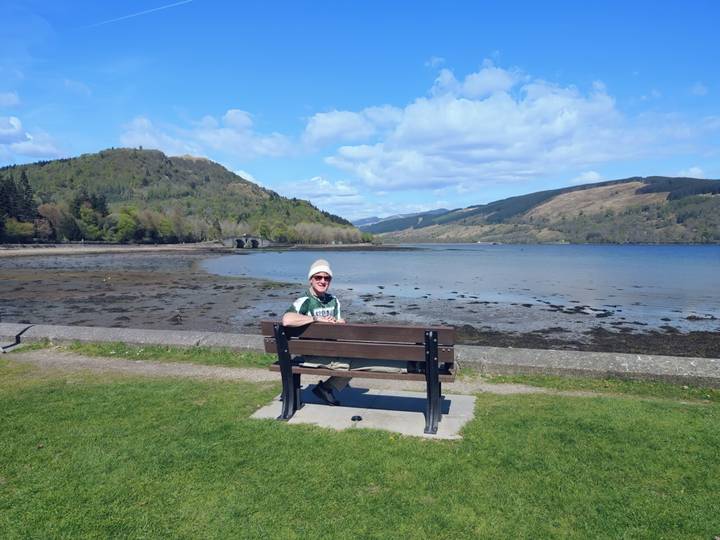 Traveler relaxing on a bench overlooking a tranquil loch and rolling green hills under blue skies.