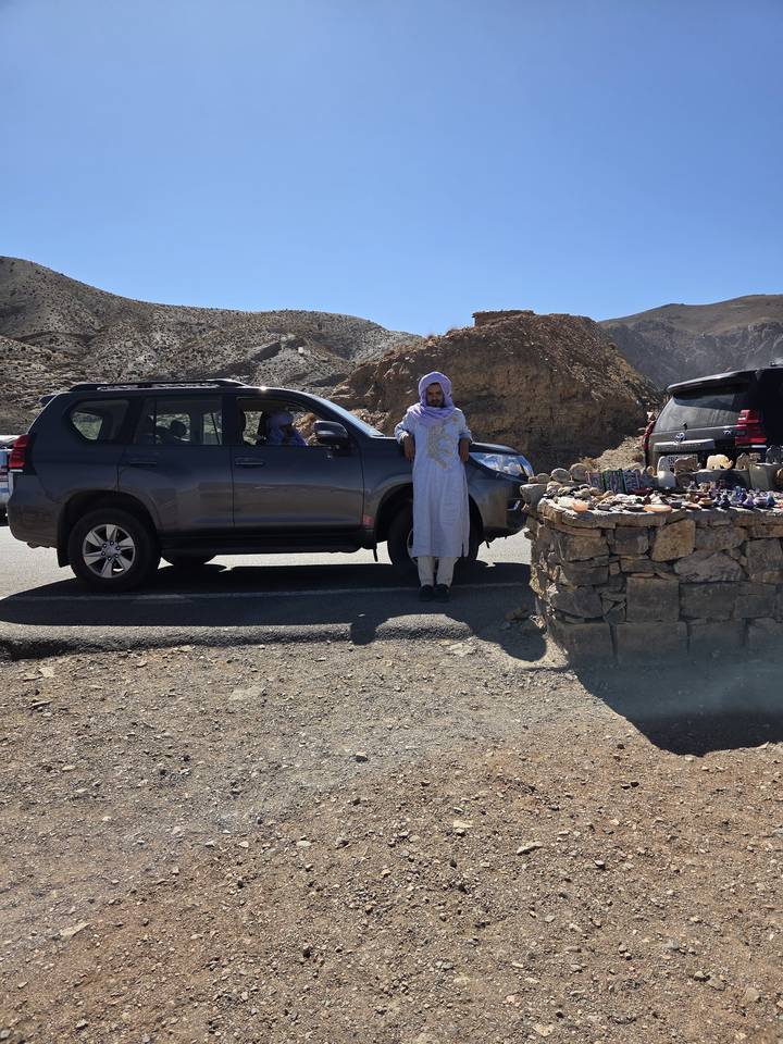 Local man in traditional robe stands beside SUV at roadside stall of stones and trinkets in rugged hills