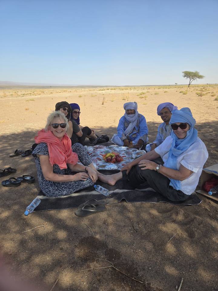 Group of travellers and Berber hosts enjoy a shaded picnic on a blanket in the desert