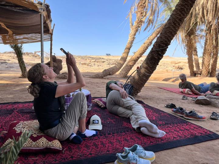 Travelers lounge on rugs under palm shade in desert camp taking photos.