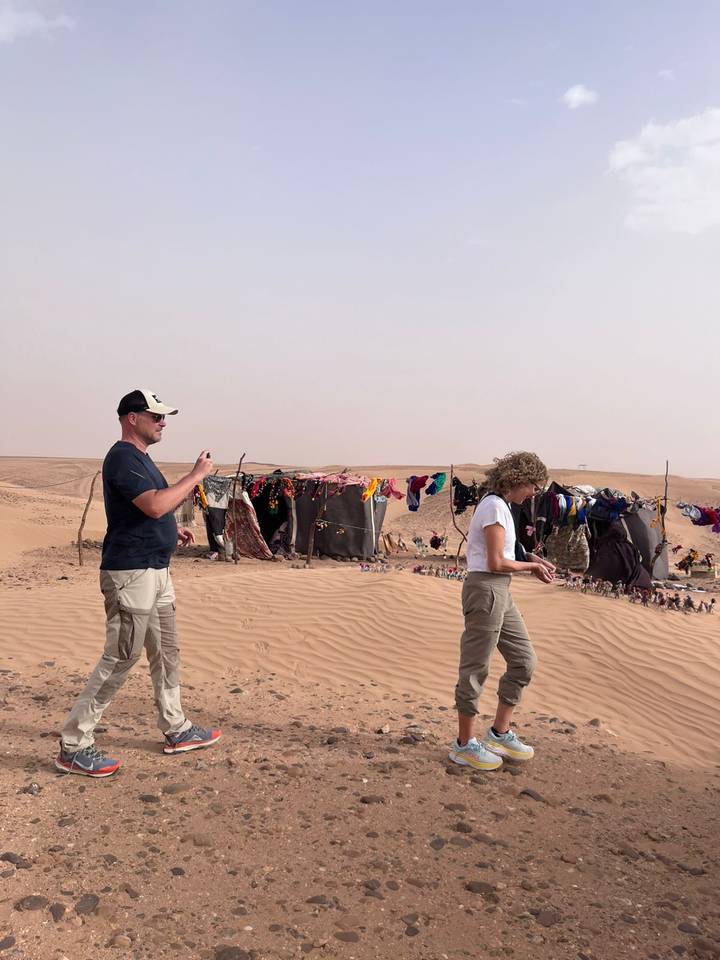 Two tourists explore a nomadic desert camp with handicrafts hanging.