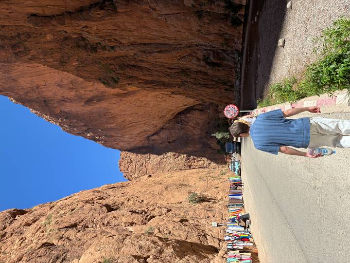 Visitor walks into the towering red limestone walls of Todra Gorge under a bright blue sky.