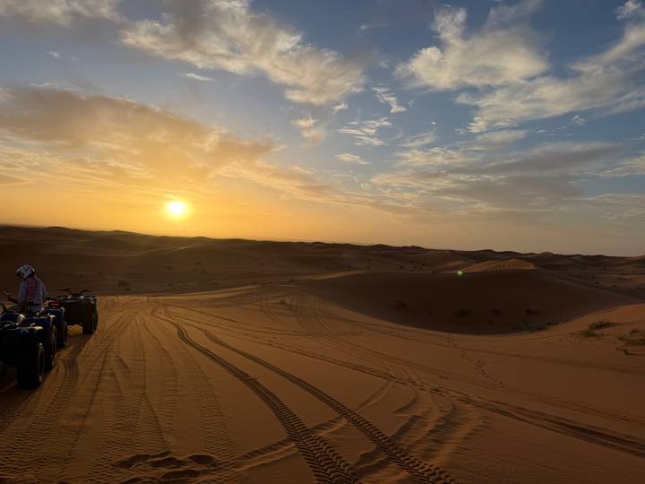 ATVs parked on sand as the sun sets over rolling Sahara dunes, casting warm orange light.